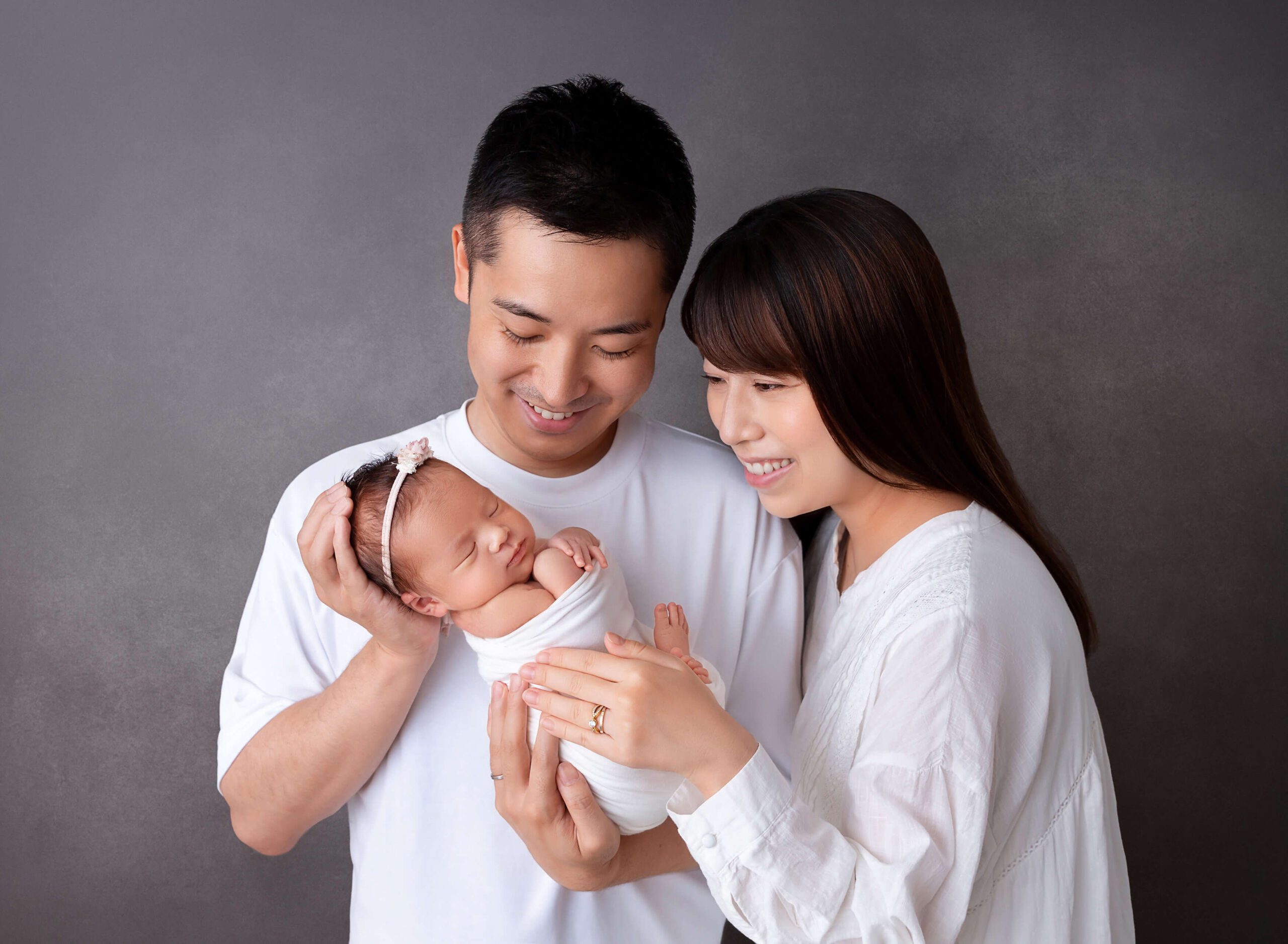 A newborn baby sleeps in mom and dad's hands while they stand in a studio in all white