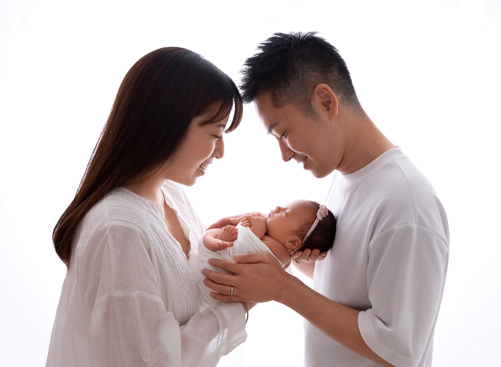 Happy new mom and dad in white smile down at their sleeping newborn daughter in their hands between them after meeting Manhattan Nannies