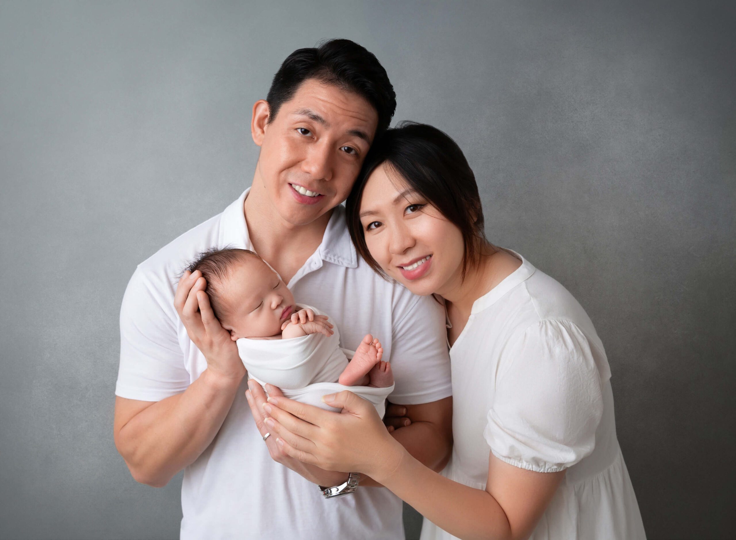 Happy new parents smile while cradling their sleeping newborn in a white swaddle after finding Manhattan Nannies