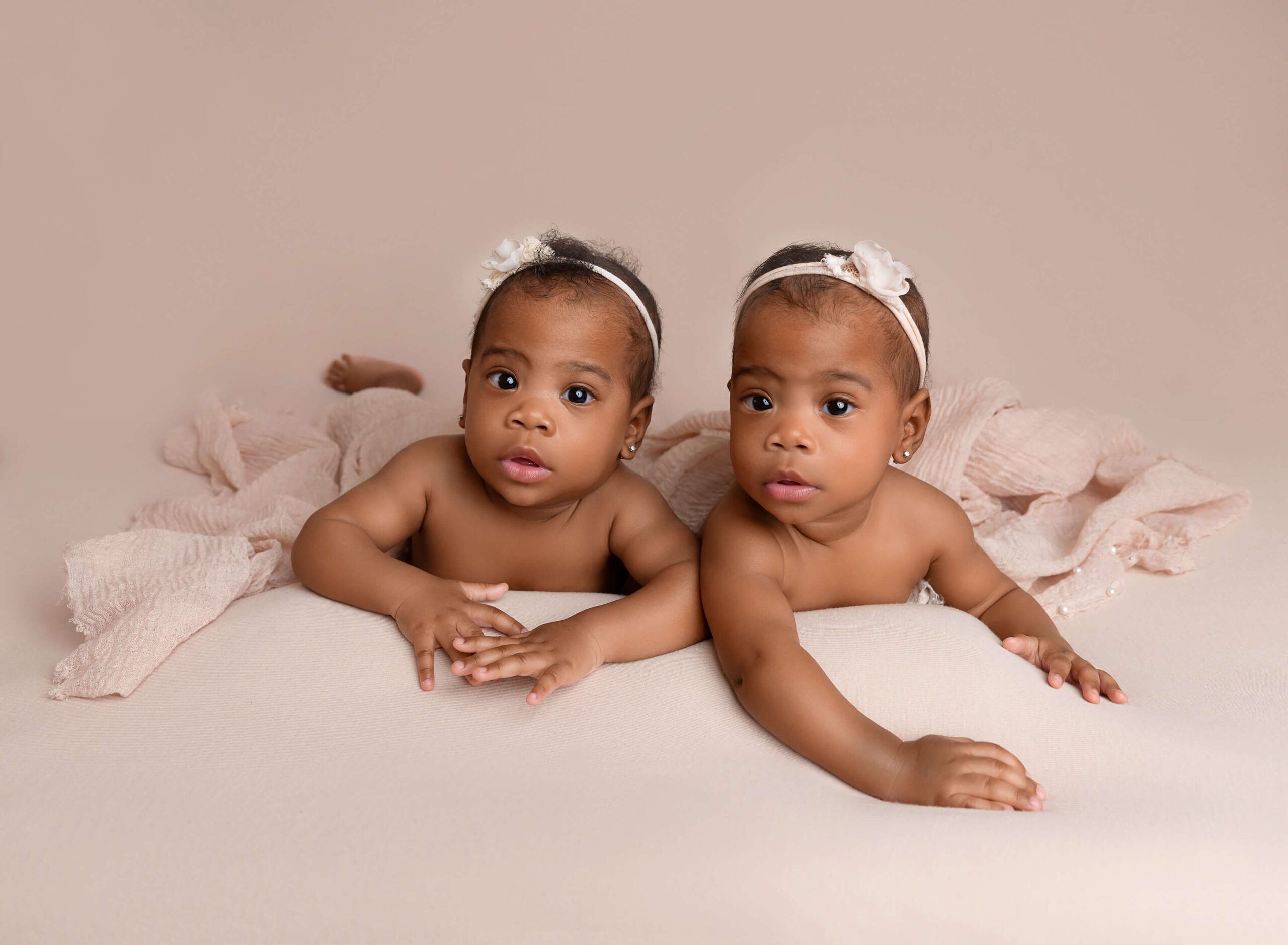 Newborn baby twin sisters lay together on a pink bed with white headbands