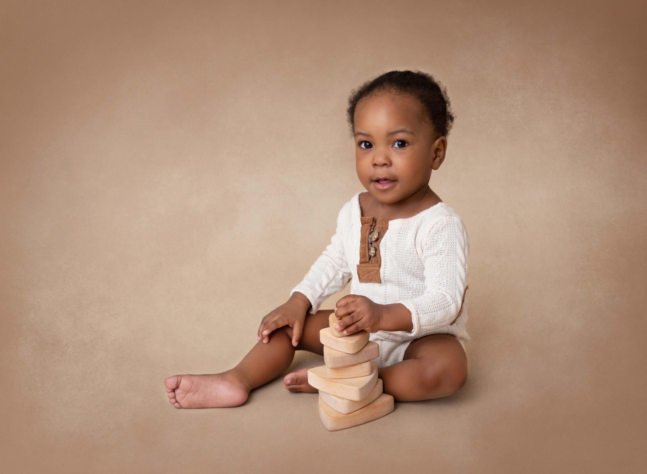 An infant sits in a white onesie playing with wooden toys in a brown studio after visiting Pediatric Chiropractors In NYC