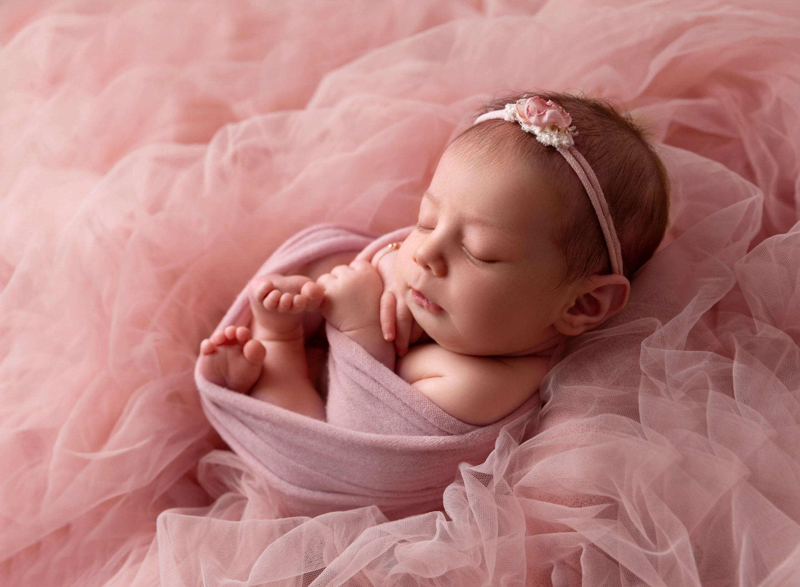 A newborn baby girl sleeps on a bed of tule in a matching pink swaddle and headband