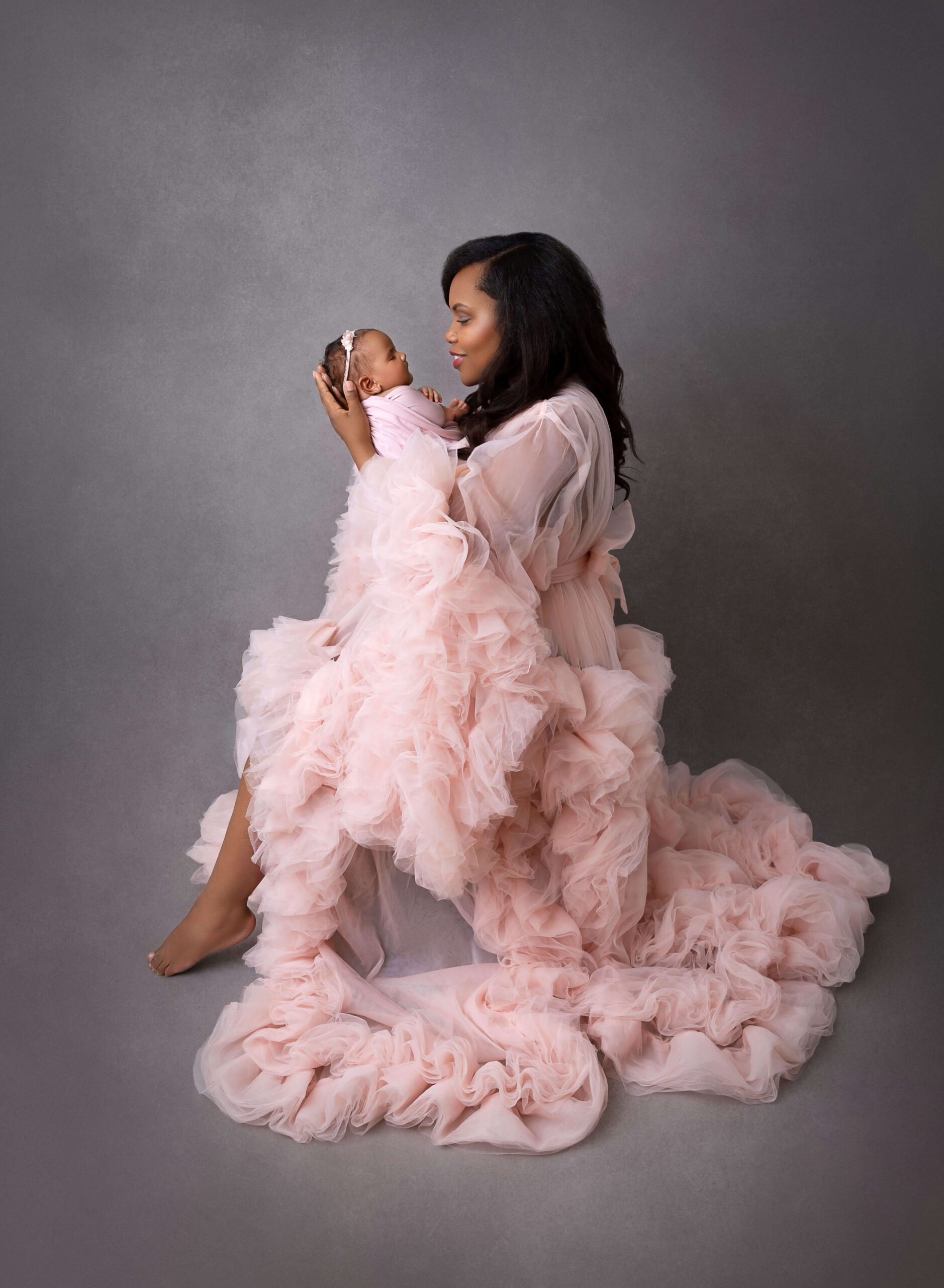 A happy mom to be sits in a flowing pink gown while cradling her newborn up to her face after visiting Posh New York