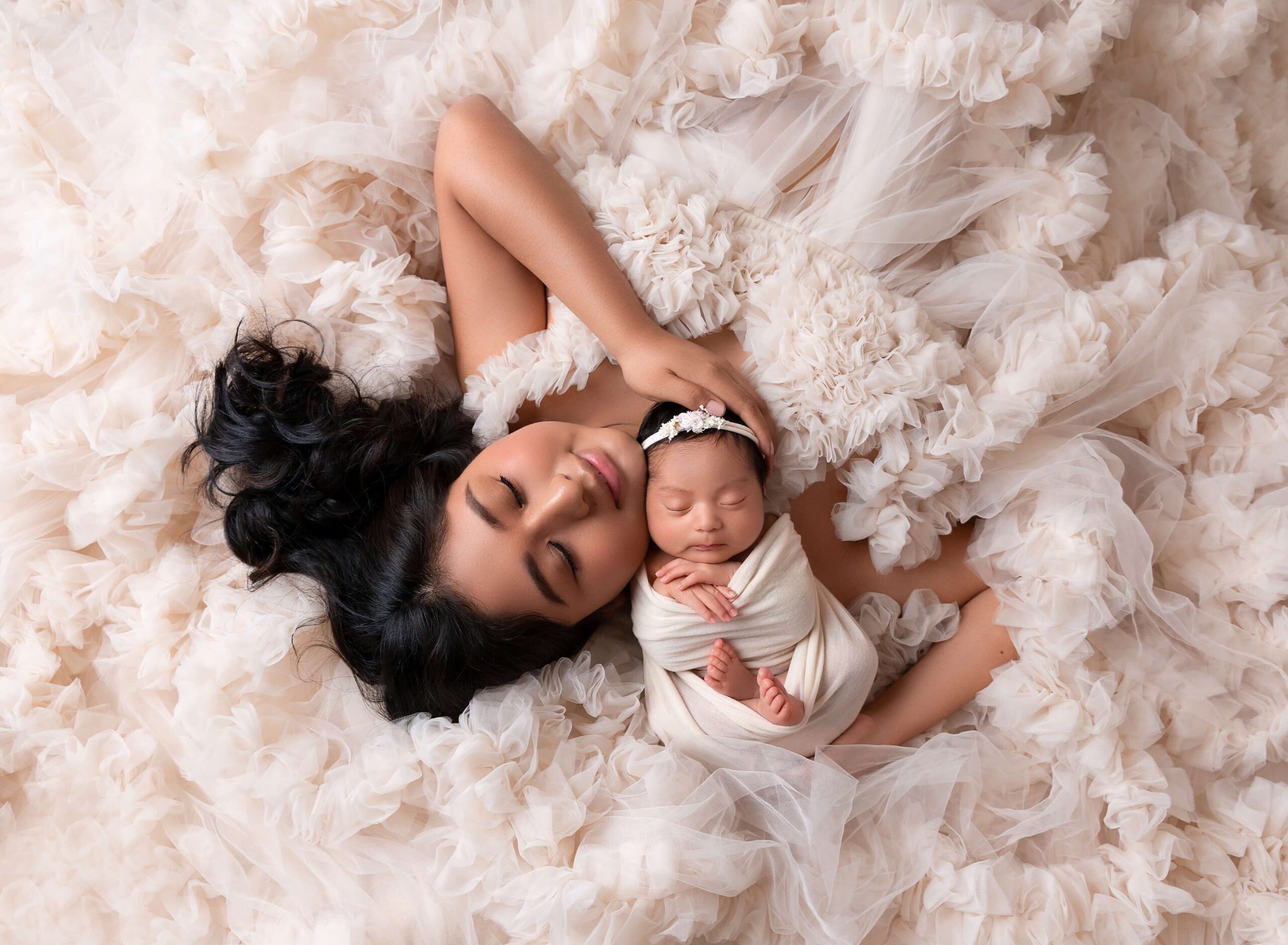 A newborn baby sleeps in a white swaddle against mom's chin on a bed of ruffled tule after visiting Posh New York