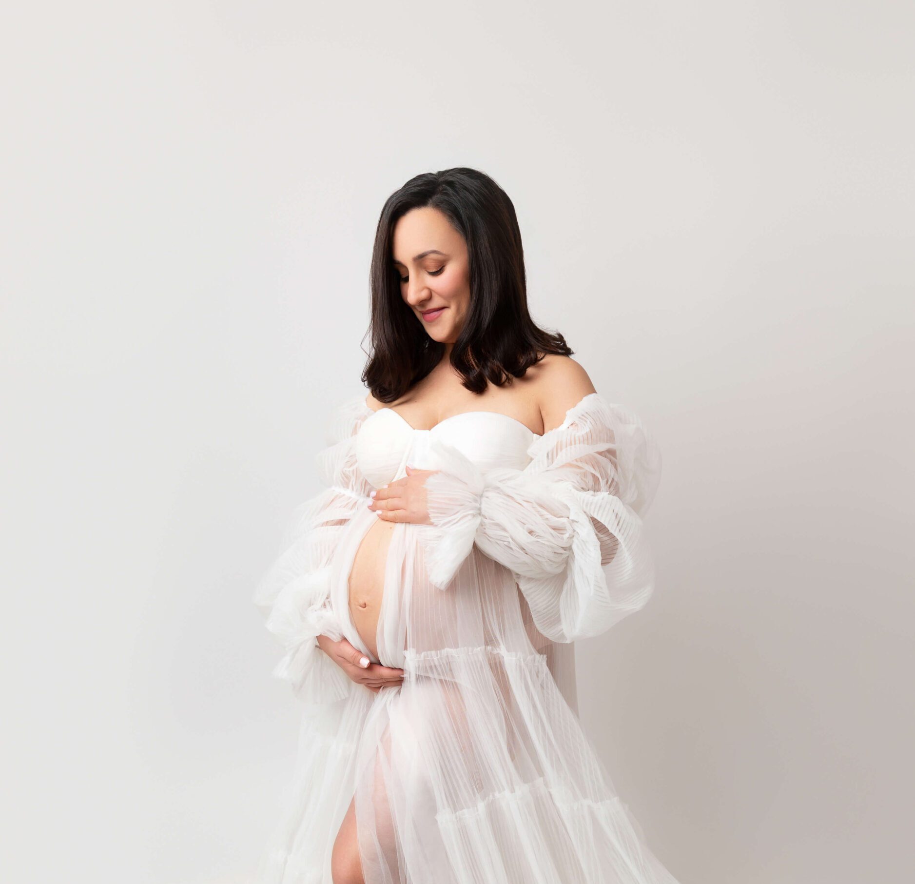A pregnant woman smiles down to her hands on her bump while standing in a studio in a white sheer maternity gown