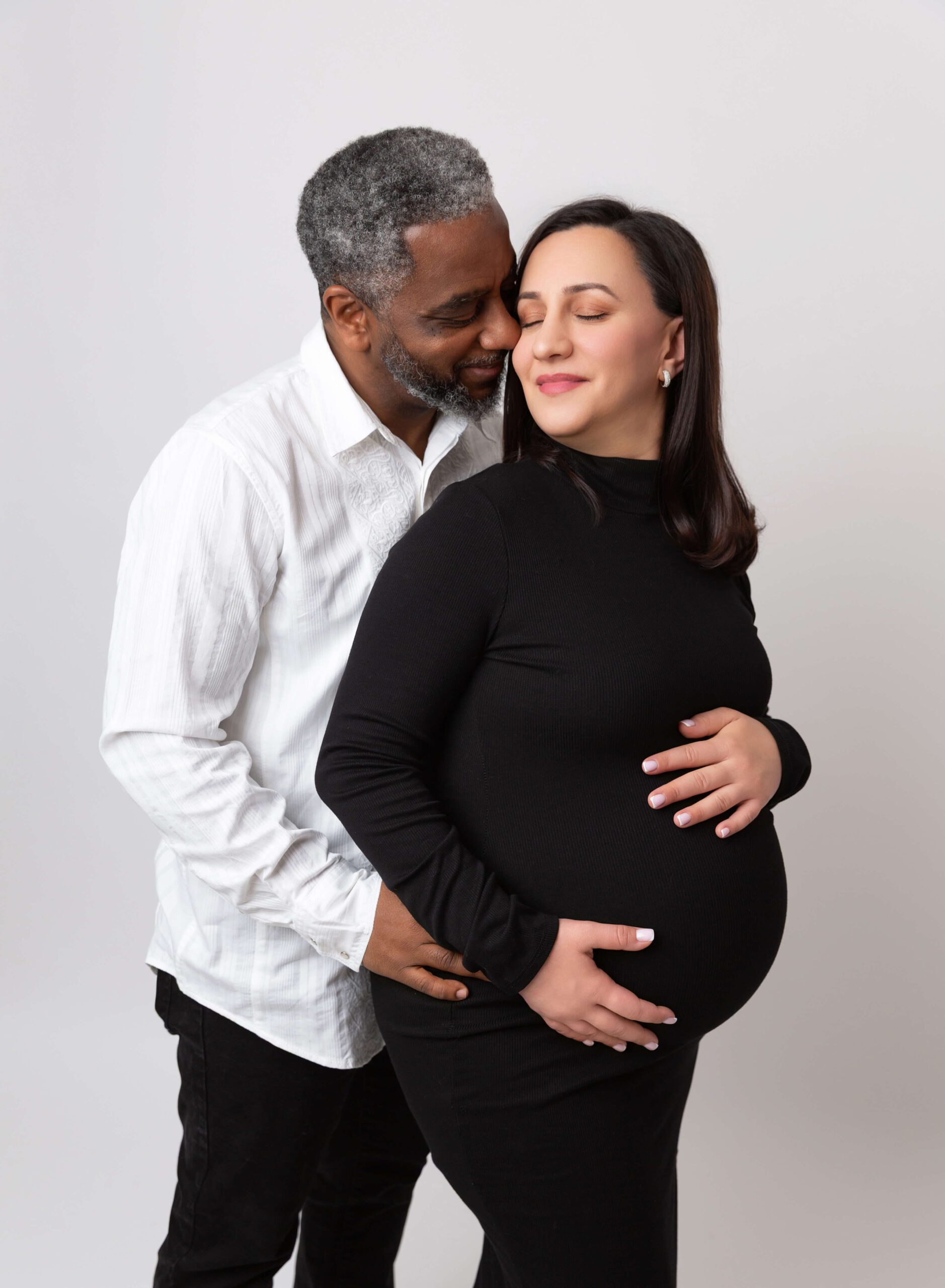 A happy expecting couple snuggles and smiles with hands on the bump in a studio after visiting Prenatal Chiropractors In NYC